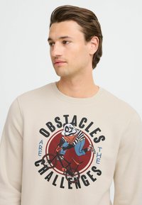 Beige sweatshirt featuring a vibrant graphic of a panda on a bicycle, text reading "OBSTACLES ARE THE CHALLENGES," round red accent background.