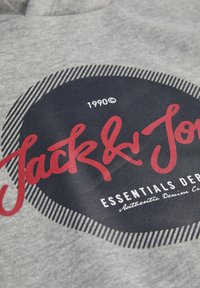 Gray sweatshirt fabric featuring a circular black logo with red text "Jack & Jones" and white accents, indicating brand and style.