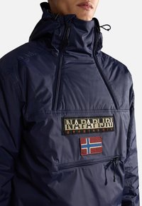 Marineblå vindjacka med hög krage, framficka med dragkedja och logopatch. Tillverkad av glänsande material och har en broderad norsk flagga.