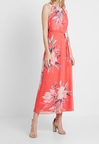 Robe maxi corail avec un décolleté halter, imprimé floral dans des teintes rose et verte, détail de taille noué et tissu fluide. Portée avec des talons neutres.