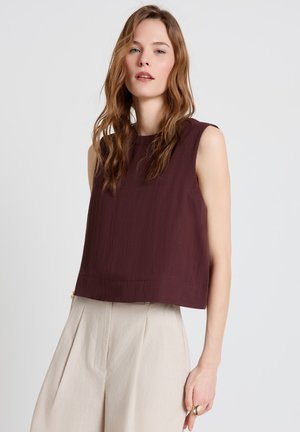 Femme aux longs cheveux ondulés châtain clair, portant un haut marron foncé sans manches et un pantalon taille haute beige clair, debout devant un fond uni.