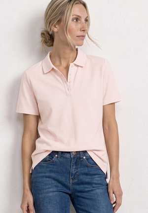 Femme aux cheveux blonds en chignon bas, portant un polo rose clair à manches courtes et un jean bleu, debout contre un mur blanc.