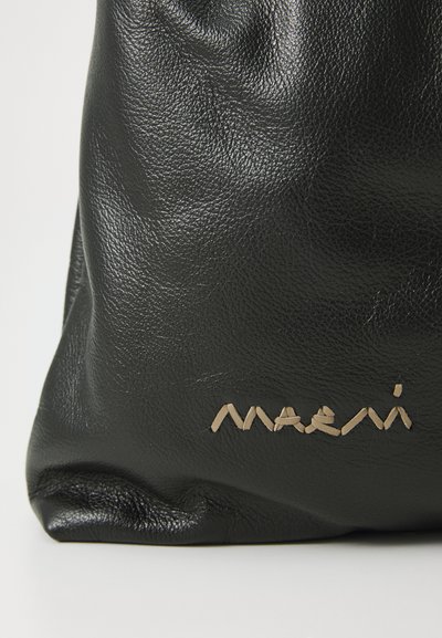 Zwarte leren tas met een textuurafwerking, met een vergulde "maeni" logo dat op de voorkant is geperst. Zachte, slungelige vorm.