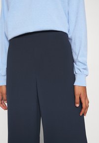 Personne portant un pantalon ample bleu marine et un haut à manches longues bleu clair, les mains détendues le long du corps sur un fond uni.