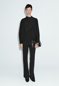 Top drappe nero con colletto alto, abbinato a pantaloni nero svasati. La modella tiene una piccola borsa a tracolla in pelle nera. Design minimalista, texture liscia.