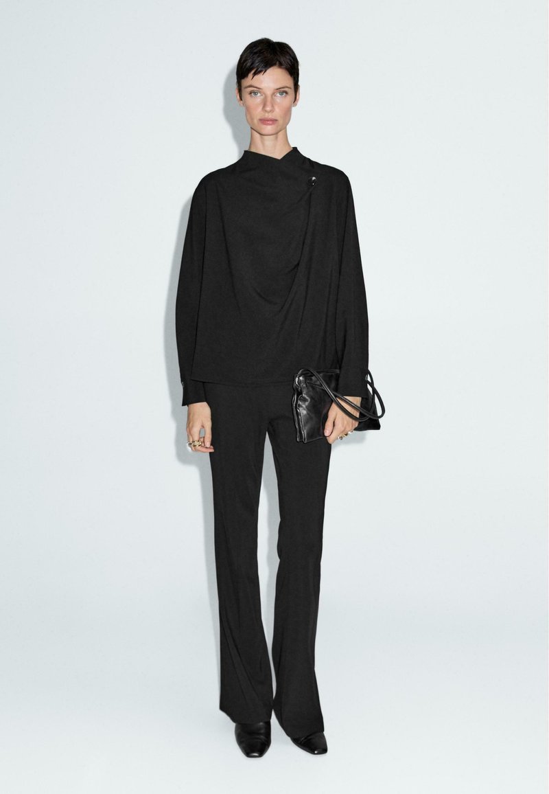 Top drappe nero con colletto alto, abbinato a pantaloni nero svasati. La modella tiene una piccola borsa a tracolla in pelle nera. Design minimalista, texture liscia.