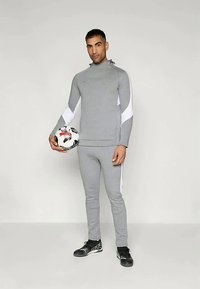 Vīrietis pelēkā sporta apģērbā tur melnbalto futbola bumbu un stāv uz vienkārša gaiša fona.