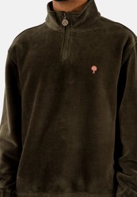 Sudadera de pana verde oliva con cuello alto, cremallera de media longitud en la parte delantera y un logo de árbol rosa en la zona izquierda del pecho. Tejido texturizado y cálido.