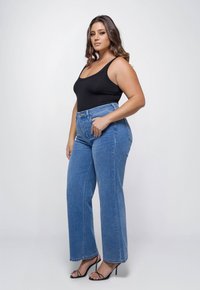 Vrouw poseert in een zwarte tanktop, hooggesneden blauwe jeans en zwarte strappy hakken tegen een plain witte achtergrond.