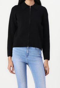 Černá zipová bunda z hladkého materiálu, s límečkem a dlouhými rukávy, v kombinaci se světle modrými přiléhavými džíny.
