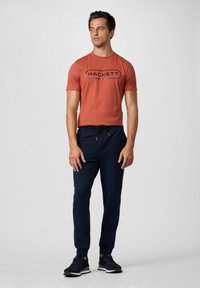 Rood-oranje t-shirt met korte mouwen met "HACKETT" grafisch ontwerp, gecombineerd met marineblauwe joggingbroek met elastische tailleband en trekkoord. Zwarte sneakers.