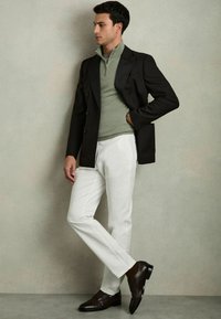 Un bărbat poartă un sacou închis la culoare, cu două rânduri de nasturi, un pulover zip-up verde deschis, o pereche de pantaloni albi și pantofi din piele maro închis.