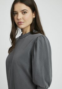 Haut gris avec un col rond et un tissu texturé. Présente des manches bouffantes avec des détails froncés. Design simple, adapté à un usage décontracté.