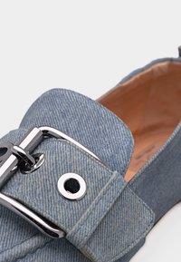 Μπλε δερμάτινο μοκασίνι με μεγάλο ασημένιο αγκράφα και λεπτομέρεια με μεταλλικά τρύπες, με ορατή εσωτερική επένδυση από δέρμα σε ταμπά απόχρωση.