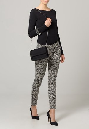 Femme portant un haut noir à manches longues, un pantalon slim à motifs, des escarpins noirs, tenant un sac à bandoulière noir avec une chaîne sur l'épaule.