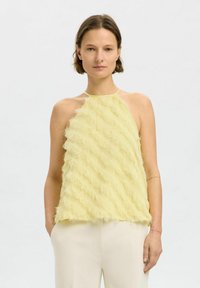 Žlutý halter top s texturovaným, třásňovým designem, se vzory připomínajícími vlny. Hladký lem a tenká ramínka. V kombinaci se světlými kalhotami.