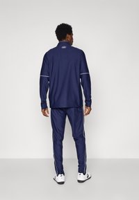 Navy atletisk treningsdrakt laget av glatt stoff, med høy krage og hvite sidelister på buksene. Sneakers med svarte såler.