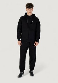 Schwarze Zip-Hoodie mit Kordelzügen, Fronttaschen und Logosiegel, kombiniert mit passenden schwarzen Jogginghosen und schwarzen Sportschuhen.