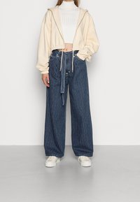 Zkrácená krémová mikina s kapucí a elastickým lemem, v kombinaci s vysokými modro pruhovanými širokými džíny a bílými teniskami.