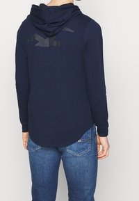 Marinblå sweatshirt med långa ärmar och avslappnad passform, med en stor svart logotyp på baksidan, mjukt tyg och vanlig nederkant.