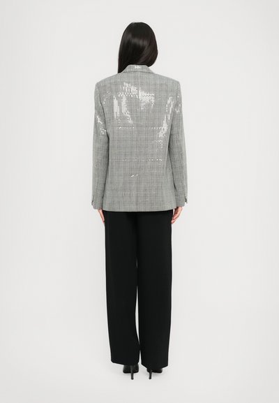 Donna in piedi con le spalle alla fotocamera, indossa un blazer a quadri grigio con paillettes, pantaloni neri a gamba larga e tacchi neri su uno sfondo bianco.