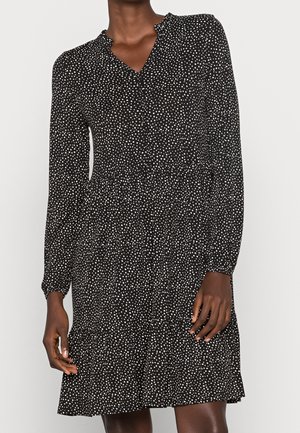 Černé šaty s dlouhým rukávem s bílými puntíky, se staženým pasem a vrstvenou sukní, nošené osobou na jednobarevném pozadí.