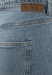 Ljusblå denimjeans med en bakficka; brun läderlogotyp sydd nära midjan med präglad text.
