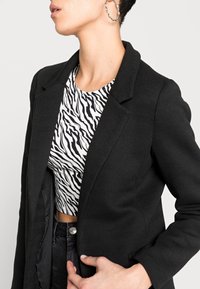 Blazer nero dal design strutturato, caratterizzato da un tessuto strutturato e rever a incavo, indossato sopra una maglia a stampa zebra bianca e nera.