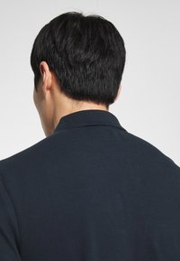 Muž s krátkými černými vlasy otočený zády, oblečený v tmavě modré košili s límečkem na jednobarevném světlém pozadí.