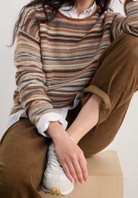 Pull rayé aux tons chauds d'orange, marron et crème. Associé à un pantalon en velours côtelé marron et des baskets blanches, assis sur un bloc de bois.