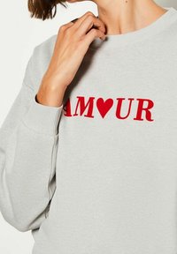 Sweat-shirt gris clair avec un texte rouge vif "AMOUR" dont la lettre "O" est remplacée par un symbole de cœur. Tissu doux et texturé.