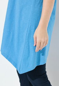 Túnica corta de manga corta en azul claro con un dobladillo curvado y un tejido suave. Una mano reposa en un lado, mostrando el ajuste relajado y la textura de la camiseta.