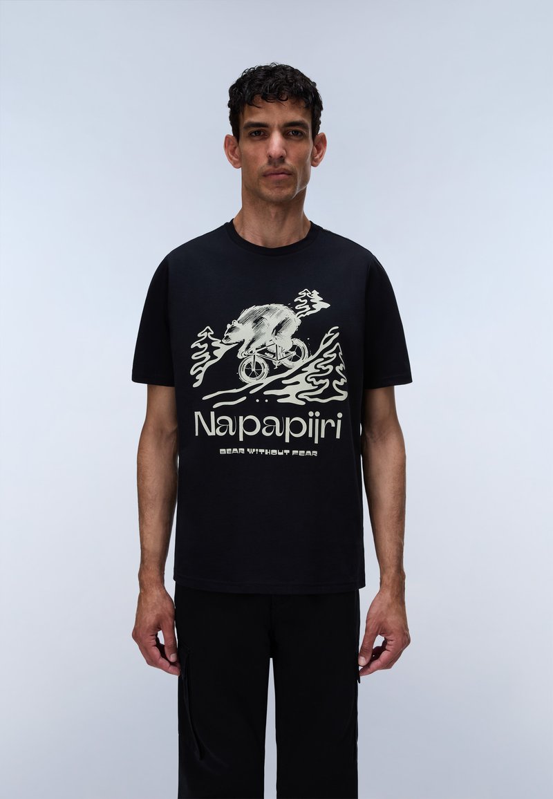 Άνδρας φοράει μαύρο T-shirt με λευκό σχέδιο αρκούδας που ποδηλατεί πάνω από κύματα, με τα κείμενα «Napapijri» και «Bear Without Fear» από κάτω.