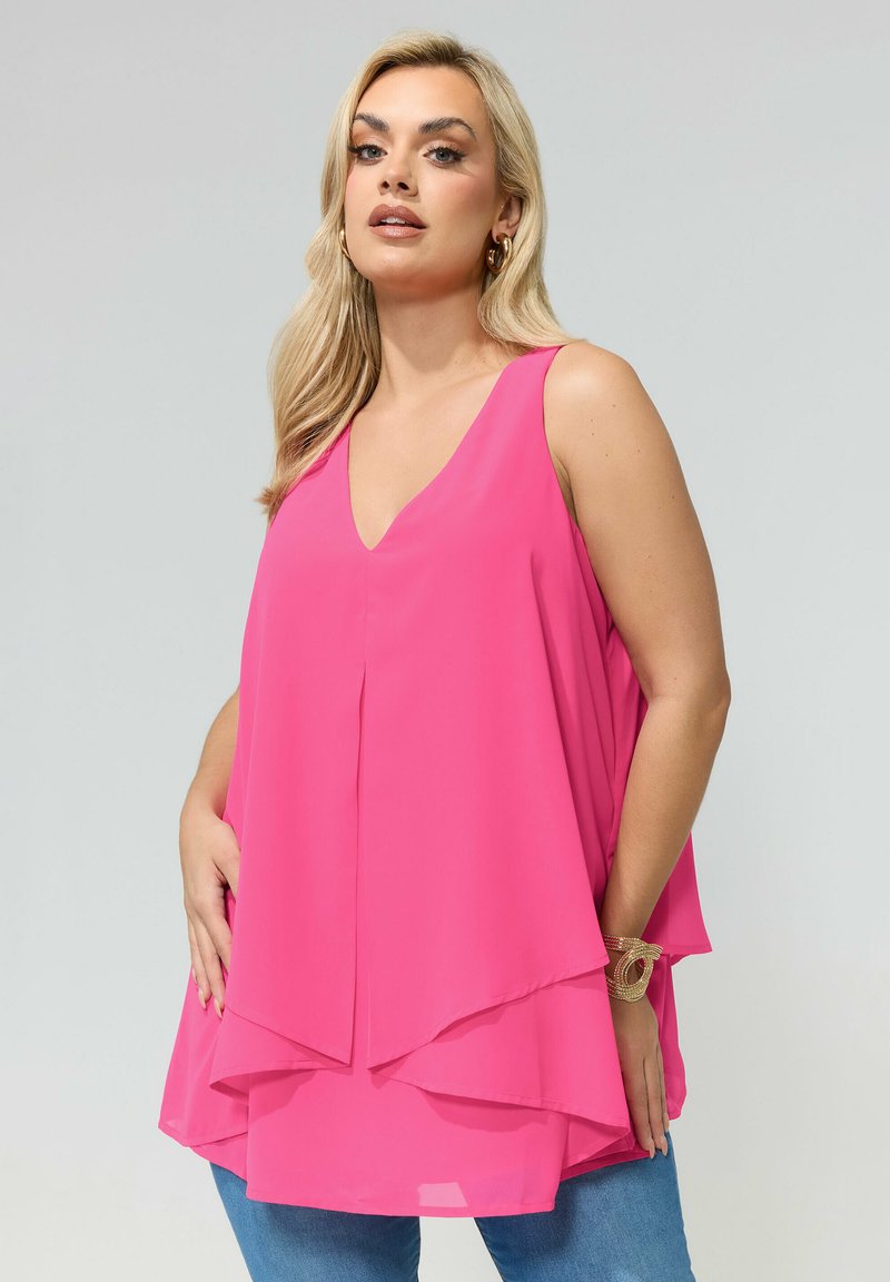 Mujer con cabello largo y rubio, lleva pendientes de aro dorados y pulsera, blusa sin mangas de capas en color rosa brillante y jeans azules, sobre un fondo claro.