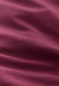 Burgundijas audums ar gludu tekstūru, kam ir smalki krunciņas un mīksta, elastīga sajūta. Tuvplāna skats izceļ tā vienmērīgo krāsu un audumu.