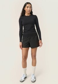 Top nero a maniche lunghe abbinato a pantaloni corti neri. Il modello indossa calze bianche e sneakers bianche con dettagli grigi. Insieme atletico semplice e casual.