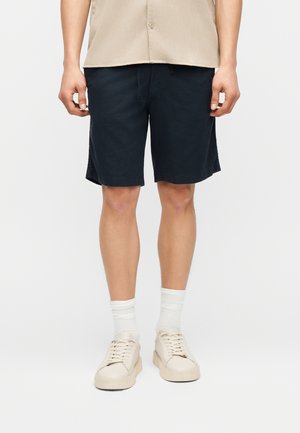 Muž v béžovej košeli, čiernych krátkych nohaviciach po kolená, bielych ponožkách po polovicu lýtok a krémových šnurovacích teniskách na jednoduchom pozadí.