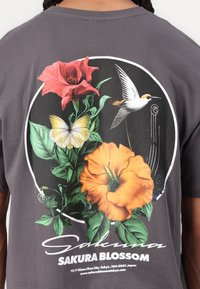 Rückseite eines grauen Hemdes mit einem kreisförmigen Design, das rote und orangefarbene Blumen, grüne Blätter, einen gelben Schmetterling und einen fliegenden weißen Kolibri zeigt.