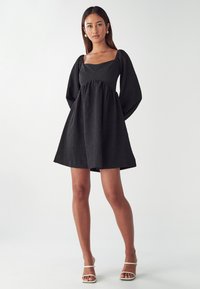 Schwarzes Kleid mit quadratischem Ausschnitt, puffigen Ärmeln, strukturiertem Stoff und taillierter Passform, das sich an der Taille weitet, kombiniert mit weißen Riemchensandalen.
