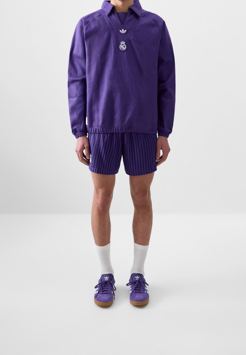 Lila långärmad topp med krage och broderad logotyp, matchad med randiga lila shorts. Bärs med vita strumpor och lila sneakers.