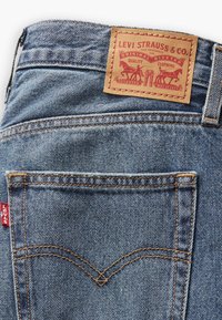 Jeans en denim bleu avec une étiquette en cuir beige, présentant des coutures rouges et une poche arrière avec un design courbé caractéristique.