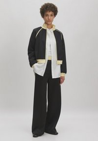 Schwarze Sportjacke mit cremefarbenen Akzenten, kombiniert mit einem weißen Hemd und schwarzen weiten Hosen. Sanfte Texturen, lockere Passform.