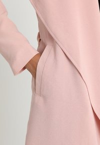 Blazer rosa chiaro con tessuto strutturato, tasche laterali e design avvolgente con spalle leggermente scese.