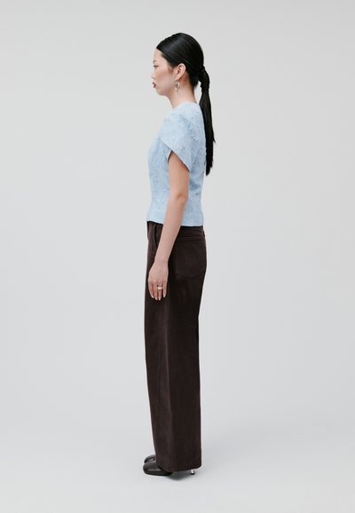 Blusa azzurro chiaro a maniche corte, con texture, abbinata a pantaloni marrone scuro a vita alta. Modella in posa di profilo contro uno sfondo neutro.