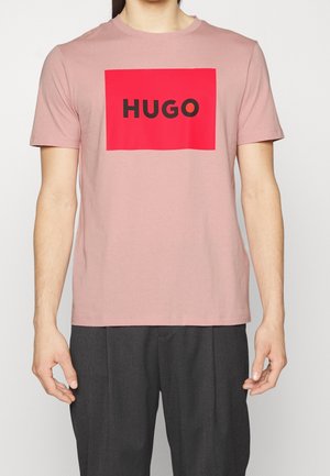 Homme portant un t-shirt rose clair à manches courtes avec un grand carré rouge et le texte noir en gras "HUGO" sur la poitrine, associé à un pantalon foncé.