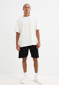 Camiseta blanca de algodón con un corte relajado, combinada con pantalones cortos negros de textura suave y diseño angular, complementada con zapatillas blancas.