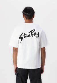 White cotton t-shirt featuring a large black "Stan Ray" logo on the back. Short sleeves, crew neck, relaxed fit. Paired with black pants.
