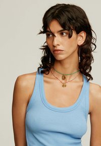 Top sin mangas azul de canalé, acompañado de dos collares en capas con colgantes de oro y acentos de cuentas. También son visibles pendientes largos con cuentas.