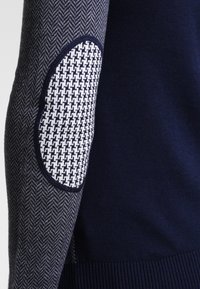 Marinblå stickad tröja med herringbone-mönstrad tyg på ärmarna och en kontrasterande armbågsfläck med ett geometriskt mönster i vitt och svart.