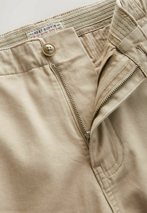 Beżowe bawełniane spodnie z zamkiem błyskawicznym, zapięciem na guzik oraz elastycznym pasem. Widoczne przeszycia wzdłuż szwów i zamka.